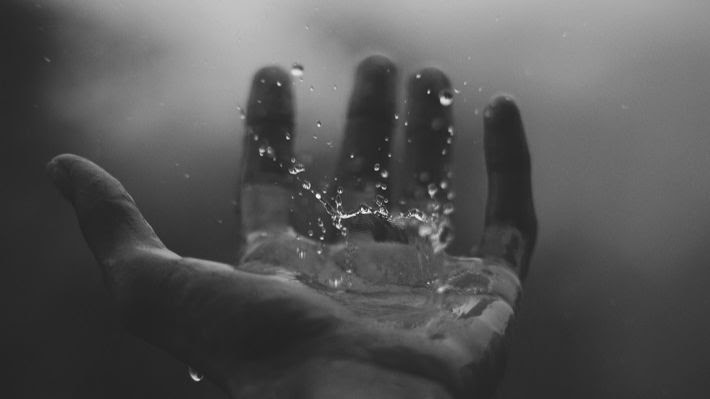 Black and white image of the left hand open from the point of view of the onlooker. There is a single drop of a clear liquid splashing on the palm of the hand. 