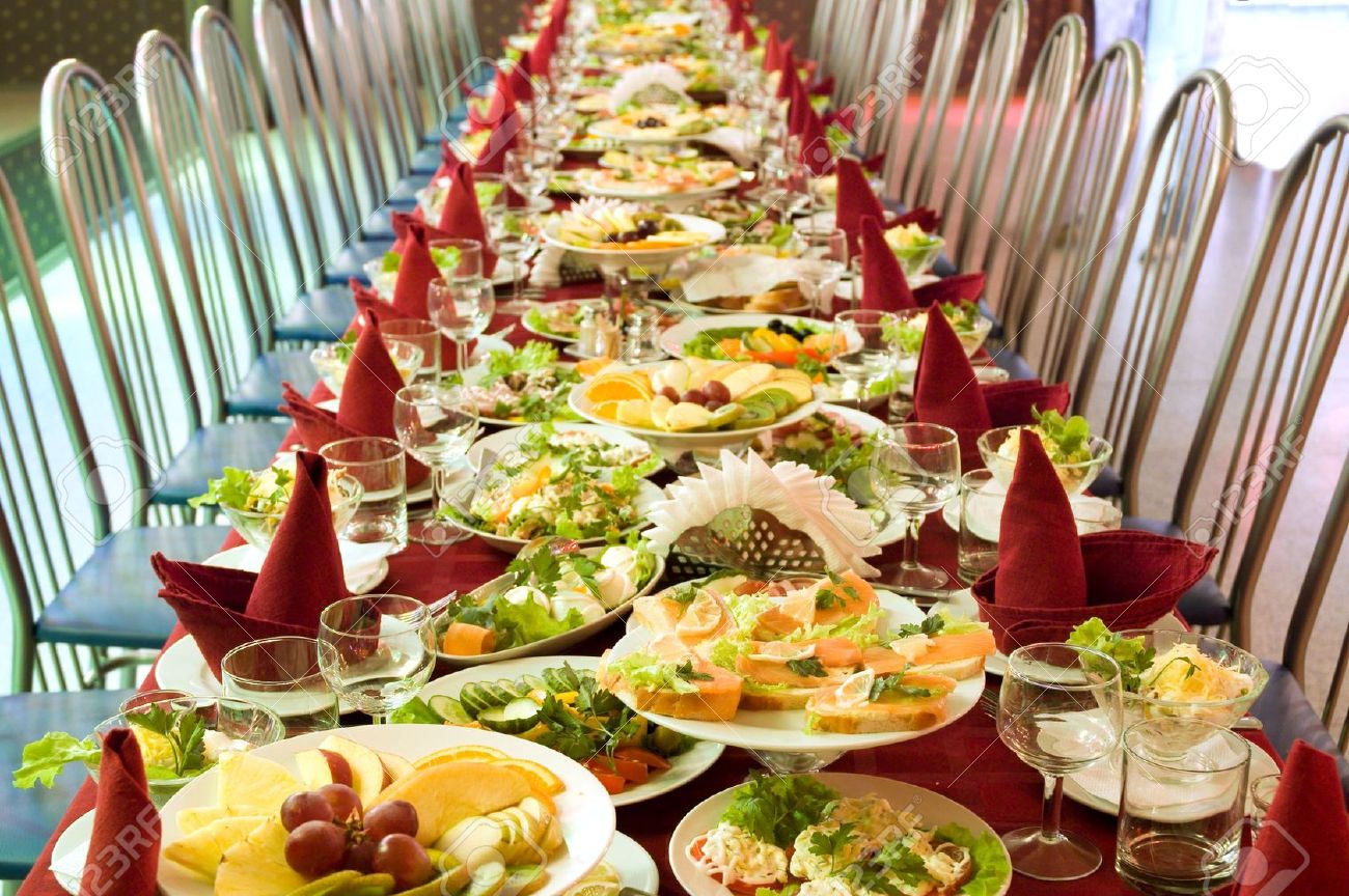 An image of a food banquet with many empty chairs. The food is primarily colorful fruits and vegetables with red napkins in the shape of birds on each plate.