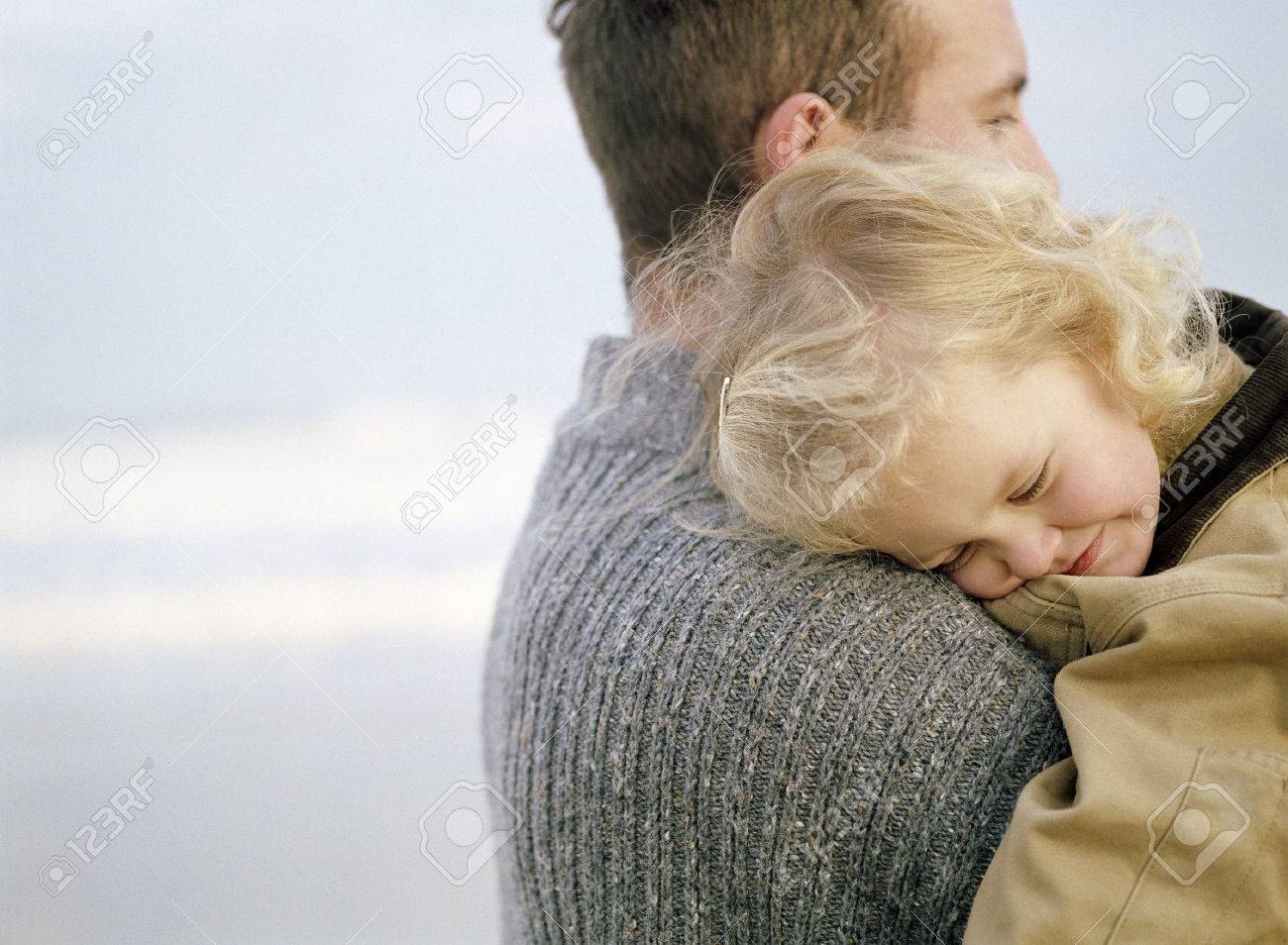 Little girl being carried on the beach by an adult male apparent figure. She has her head on his shoulder.