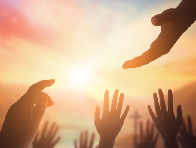 The image is of one hand reaching down to multiple hands reaching up. In the background is a multi colored sunset and a cross in the distance.