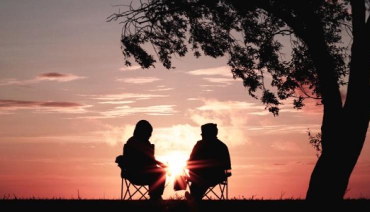 An image of the silhouette of two people sitting and talking calmly in front of an orange hued sunset. There is a tree in the foreground.